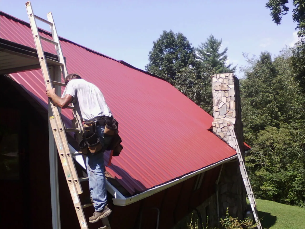 Roofer on a ladder beside a red metal roof and stone chimney — Soffit & Fascia Repair by Roofing Brothers in Ranger